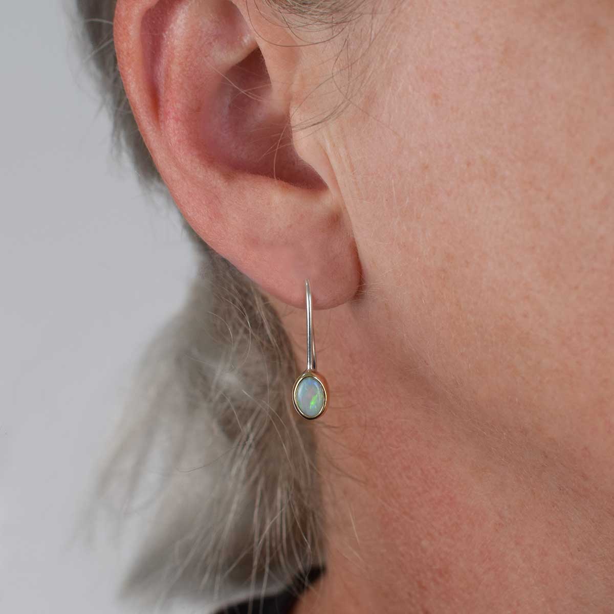 A close-up of a person's ear wearing a small drop earring with a silver-toned hook and an oval iridescent stone in a gold-toned bezel setting.