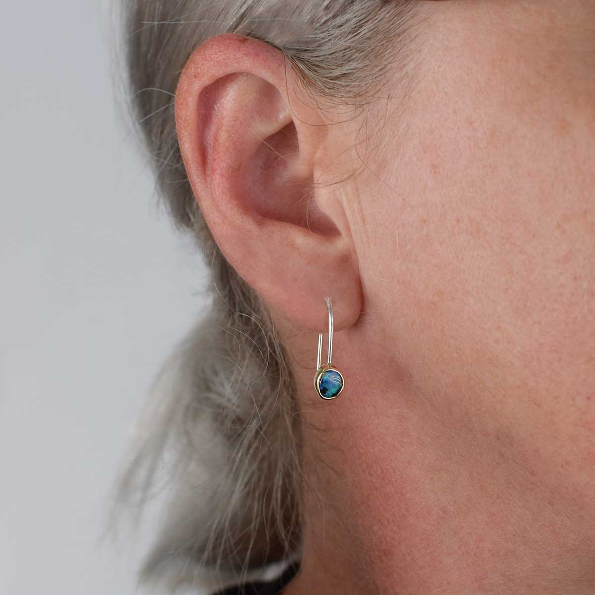A close-up of a person's ear wearing a delicate silver huggie hoop earring with a small oval blue stone in a bezel setting.
